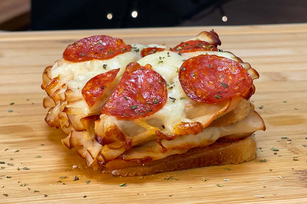Side view of a high-protein pizza grilled cheese sandwich with layers of deli chicken slices, melted mozzarella cheese, and pepperoni on a wooden cutting board.