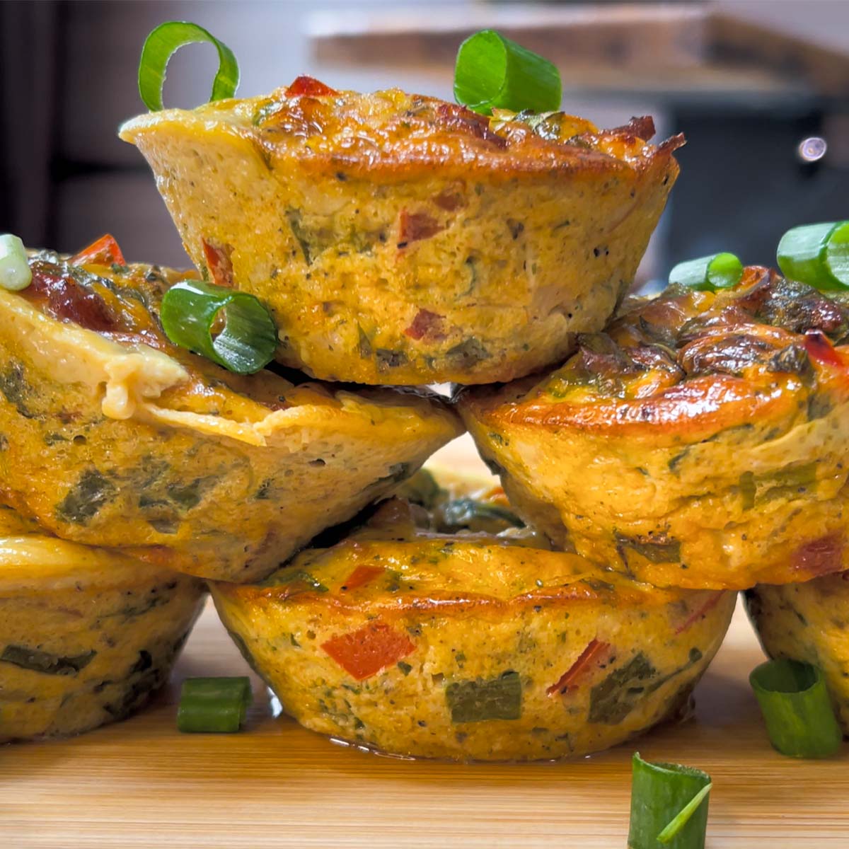 Close-up stack of baked egg bites on a wooden board, garnished with chopped green spring onions.