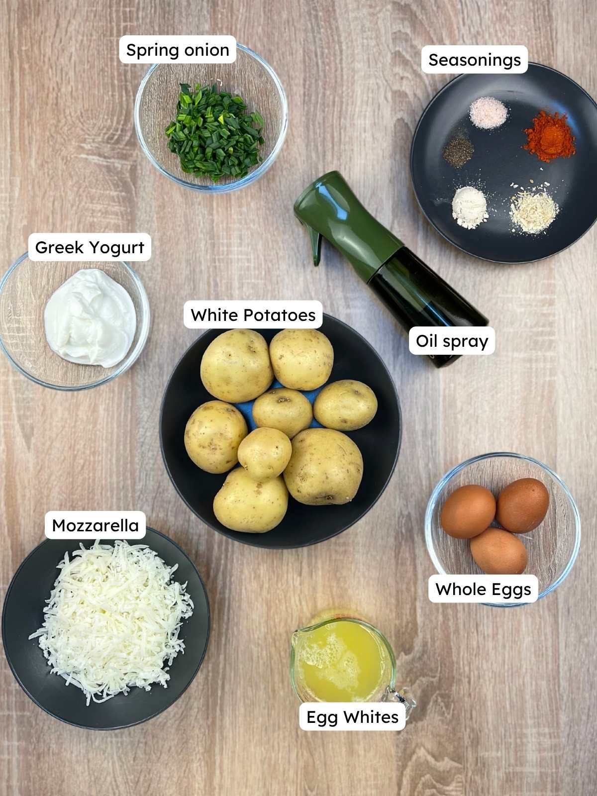 Labeled ingredient overview on a wooden table showing everything needed to make the high-protein breakfast meal prep bowls, including white potatoes, whole eggs, egg whites, Greek yogurt, mozzarella, spring onions, seasonings, and oil spray.