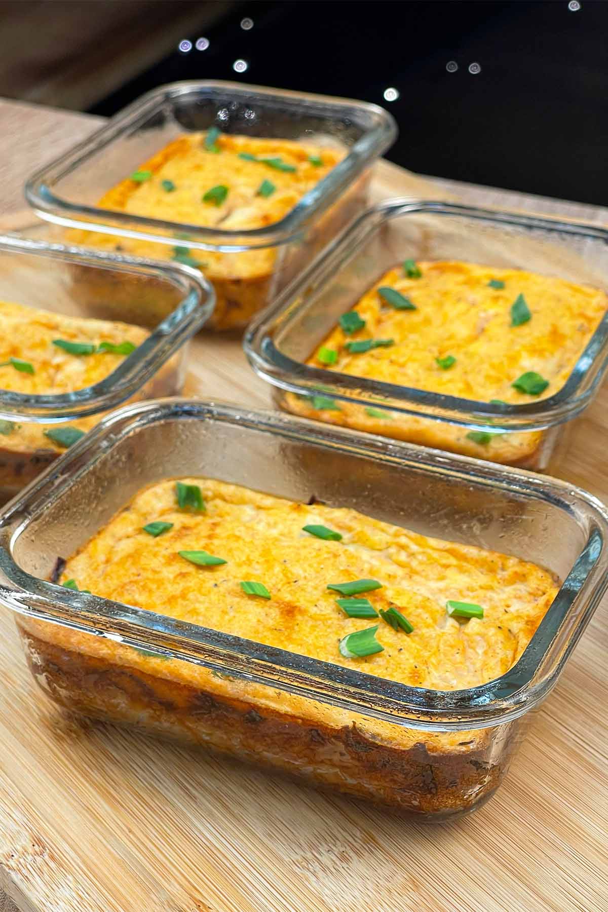 Vertical portrait shot of four finished high-protein breakfast meal prep bowls lined up in glass containers on a wooden cutting board.
