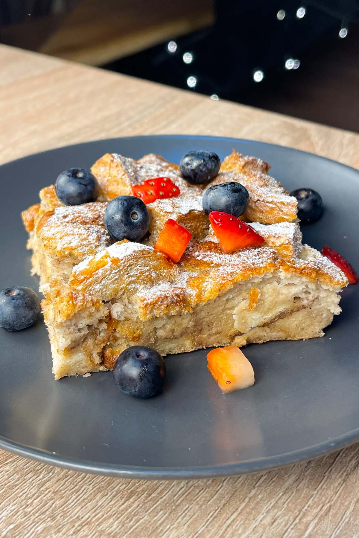 Side view of a slice of baked high-protein French toast casserole served on a dark gray plate, dusted with powdered sweetener and topped with fresh blueberries and chopped strawberries.