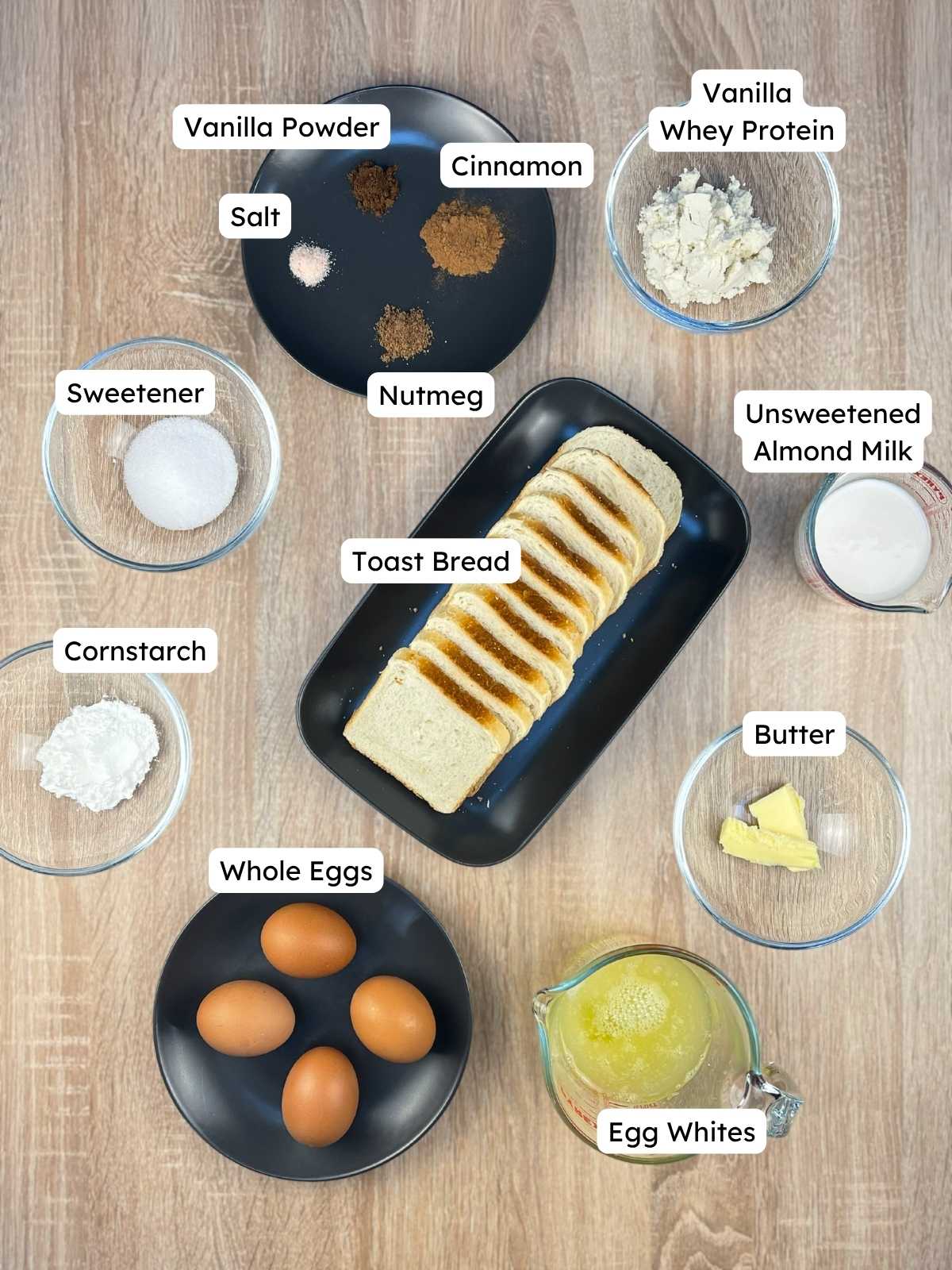 Labeled ingredient overview on a wooden table showing everything needed to make the high-protein French toast casserole, including toast bread, egg whites, whole eggs, vanilla protein powder, almond milk, and spices.