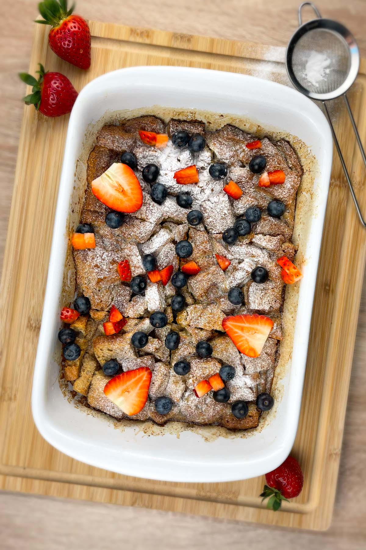 Vertical top-down shot of the finished high-protein French toast casserole in its baking dish, surrounded by whole strawberries and a powdered sugar sifter.