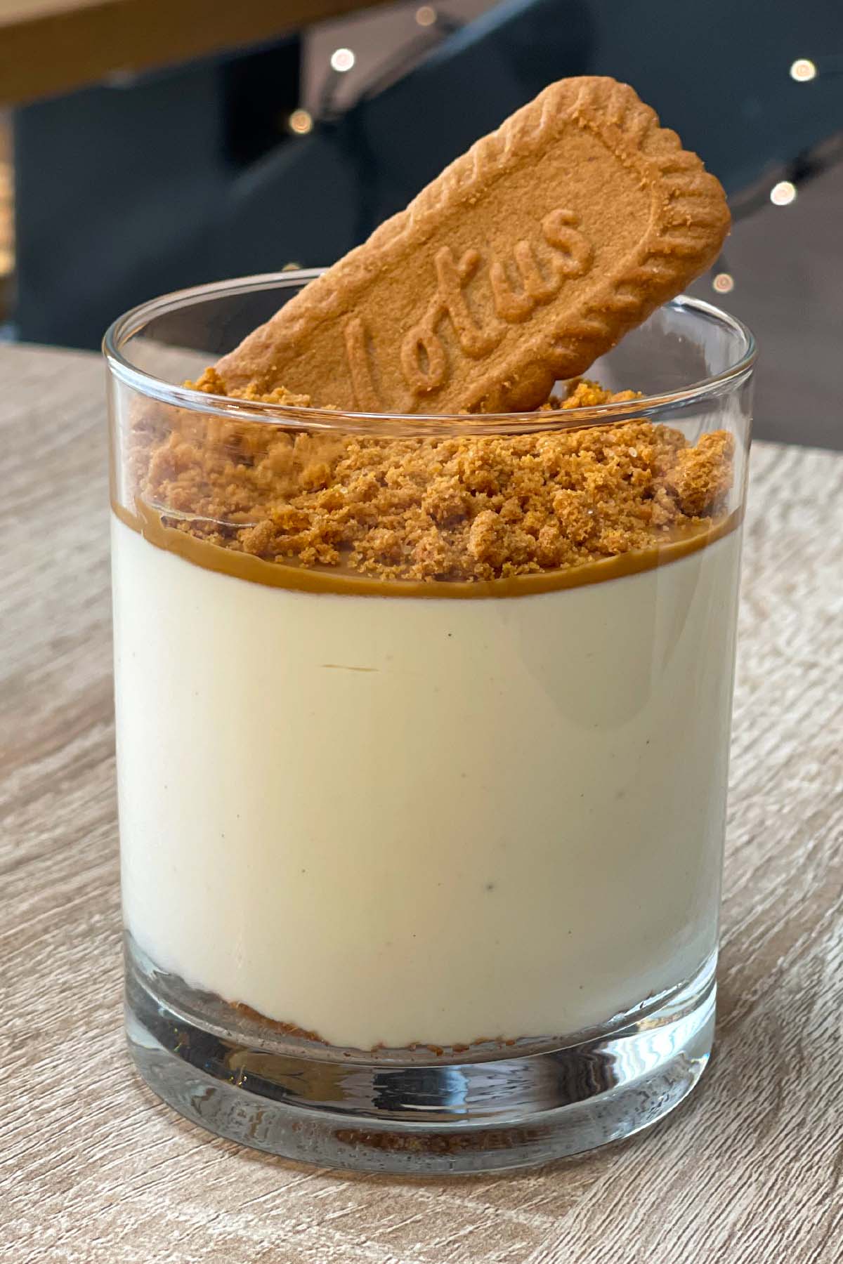 Vertical close-up of a high-protein Biscoff cheesecake cup layered with a biscuit base, cheesecake filling, cookie butter spread, crushed biscuits, and a whole Lotus Biscoff cookie.