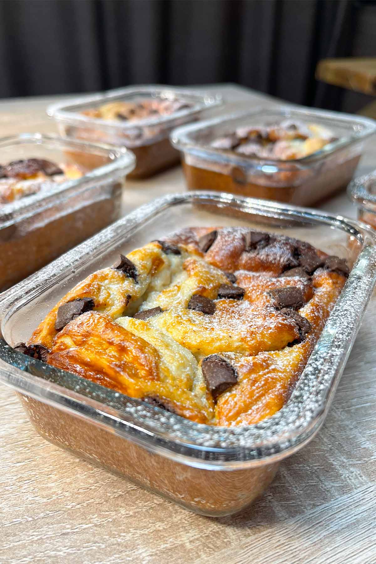 Close-up of a baked chocolate chip protein pancake bowl in a glass container, dusted with powdered sugar on a wooden table.