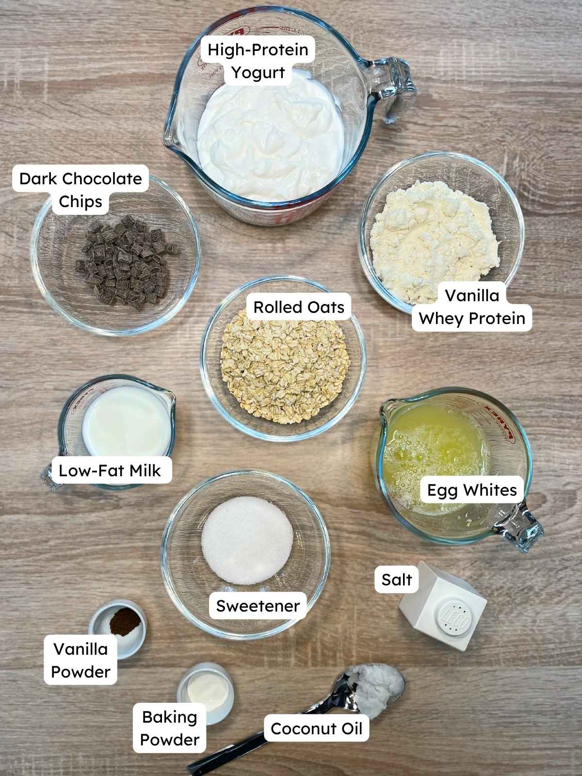 Labeled ingredient overview on a wooden table showing everything needed to make protein pancake bowls, including yogurt, oats, protein powder, egg whites, and chocolate chips.