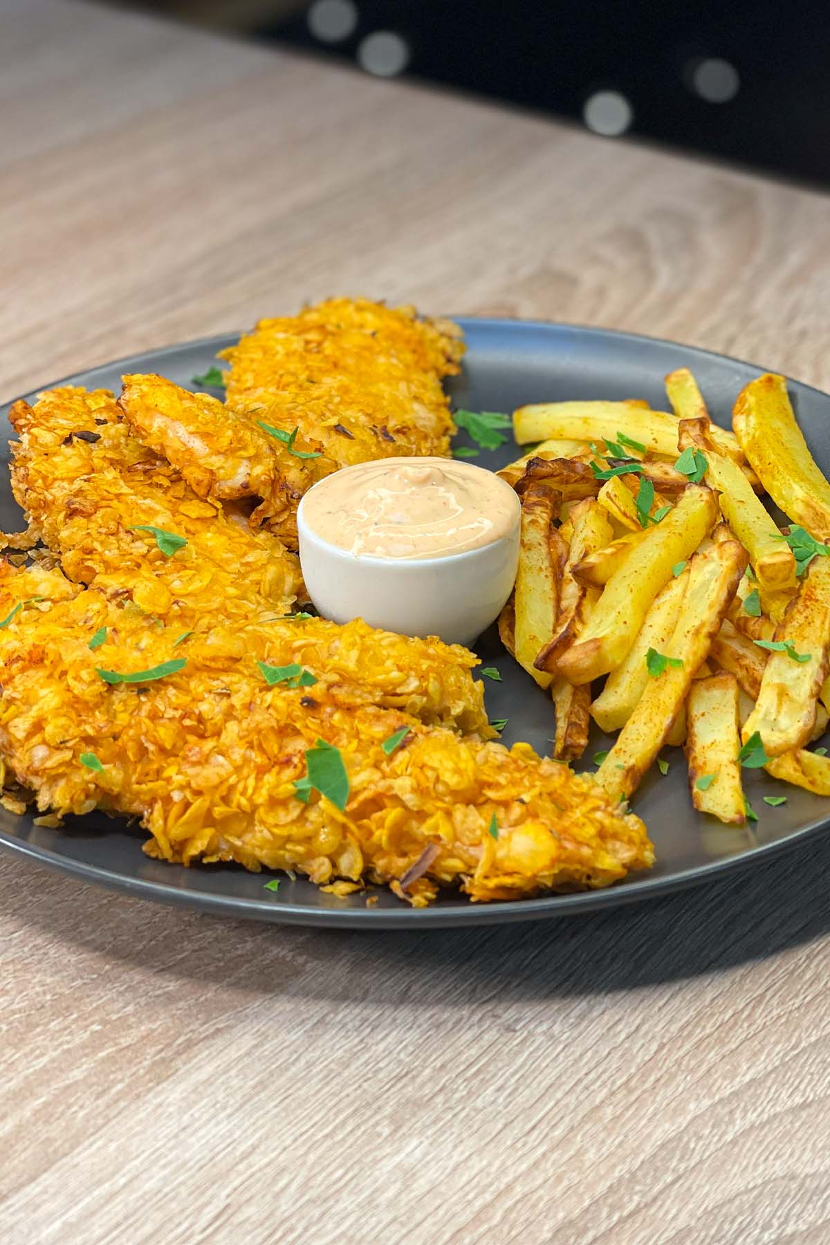 Side view of golden-brown air fryer chicken strips coated in crispy cornflakes, served on a dark plate with homemade french fries and a small bowl of dipping sauce.