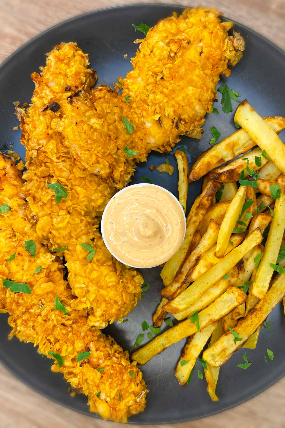 Top-down view of a plate with crunchy cornflake chicken strips, golden french fries garnished with fresh parsley, and a cup of dipping sauce in the center.