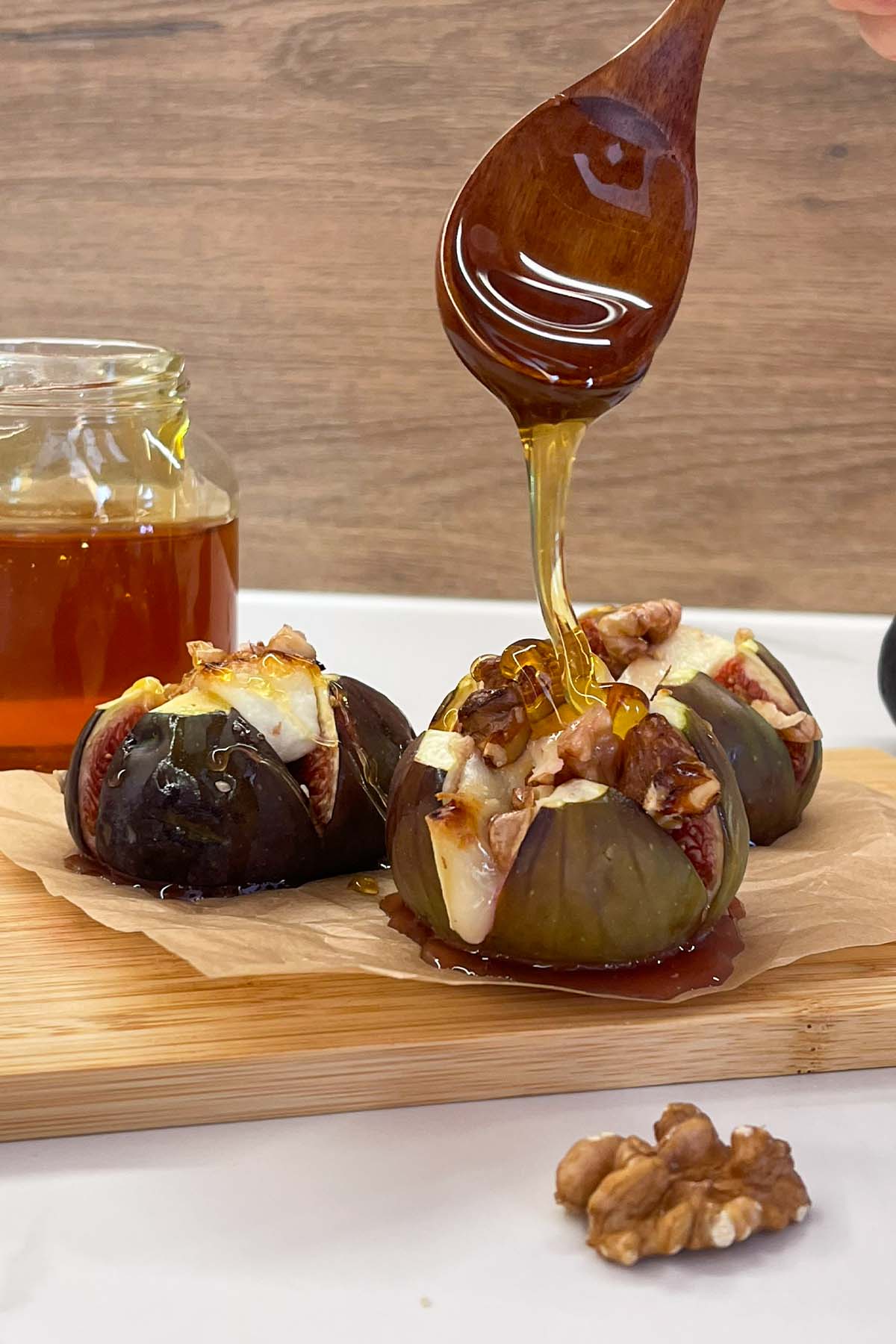 Close-up of honey being drizzled from a wooden spoon onto warm baked figs stuffed with melted goat cheese and walnuts on a wooden board.