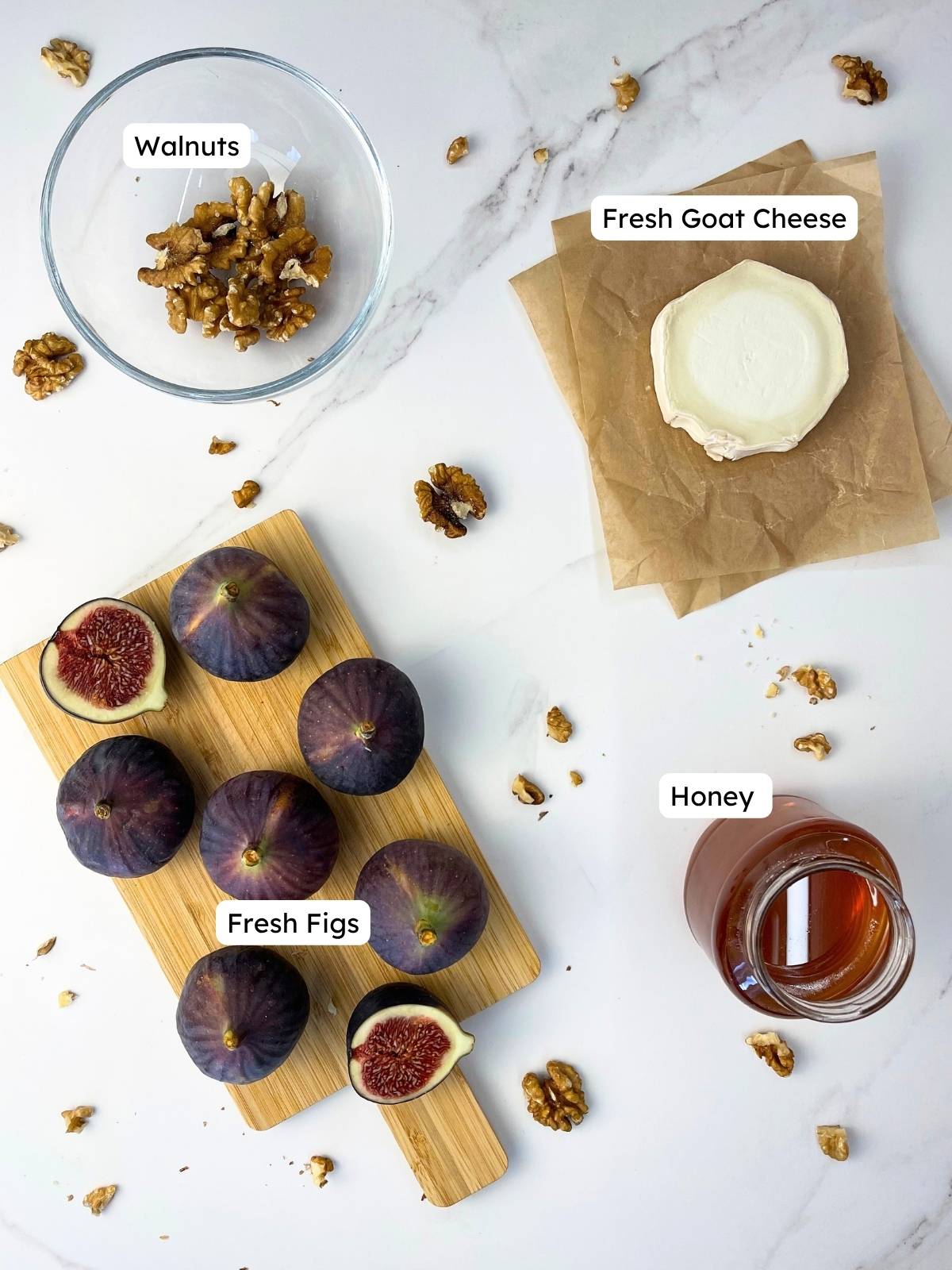 Labeled ingredient overview on a white marble background showing everything needed to make baked figs with goat cheese, including fresh figs, a round of fresh goat cheese, walnuts, and honey.