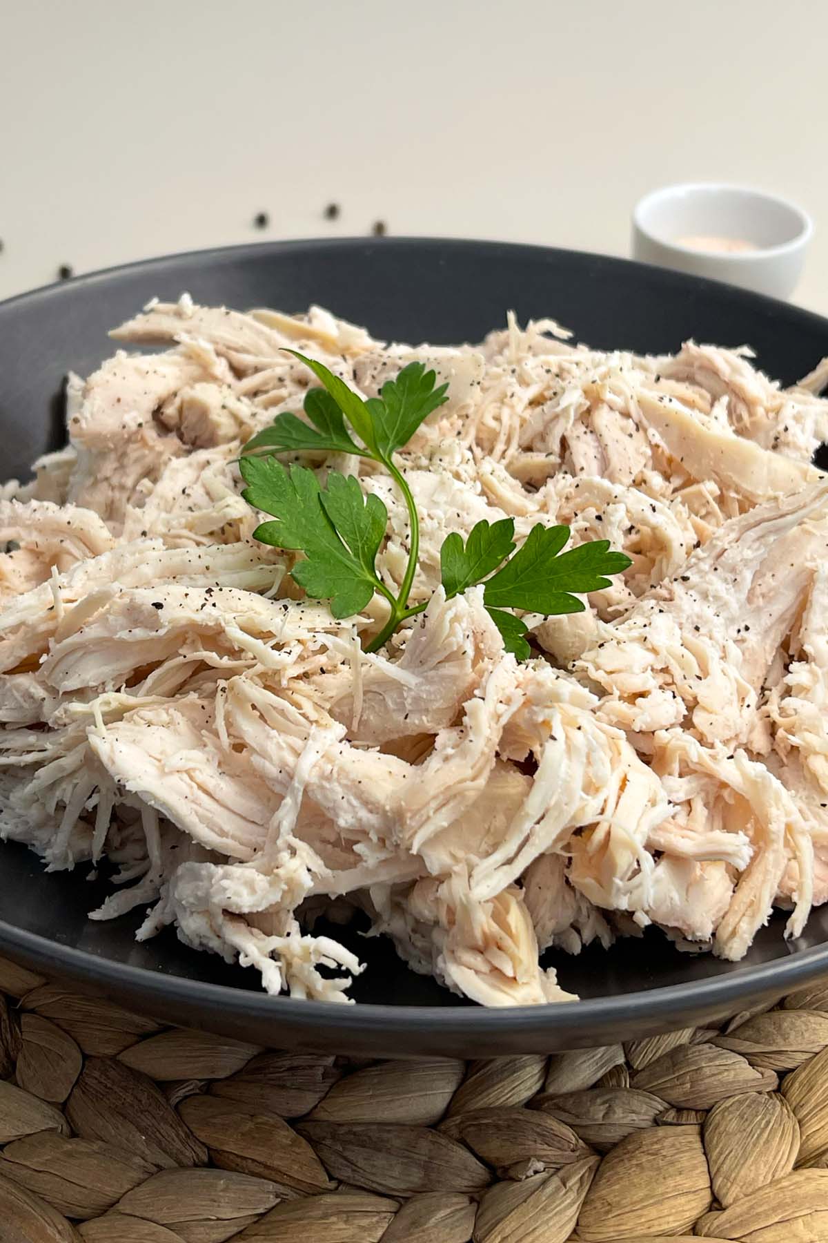 Close-up shot of a dark bowl piled high with juicy shredded chicken, seasoned with cracked black pepper and garnished with fresh parsley.