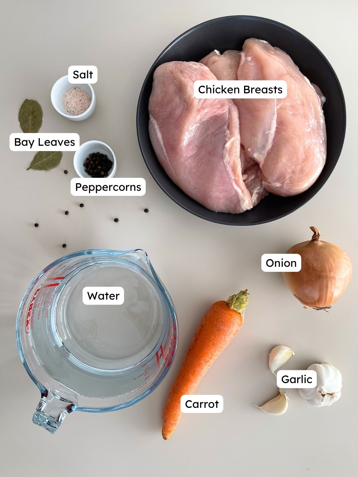 Labeled ingredient overview on a clean background showing everything needed to make shredded chicken, including raw chicken breasts, water, carrot, onion, garlic cloves, bay leaves, peppercorns, and salt.