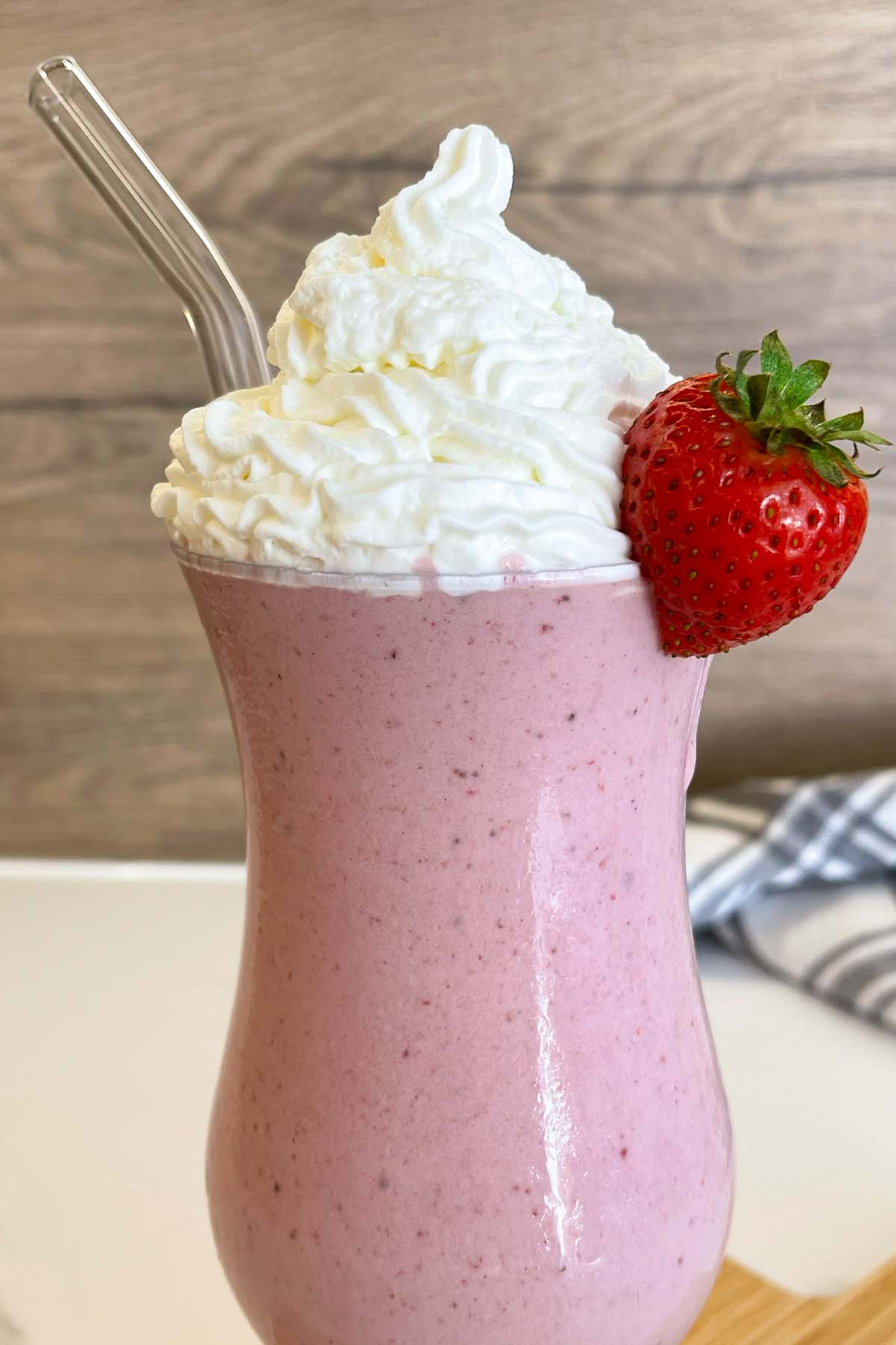 Close-up shot of a creamy protein strawberry milkshake in a tall glass, topped with light whipped cream, a fresh strawberry, and a glass straw.