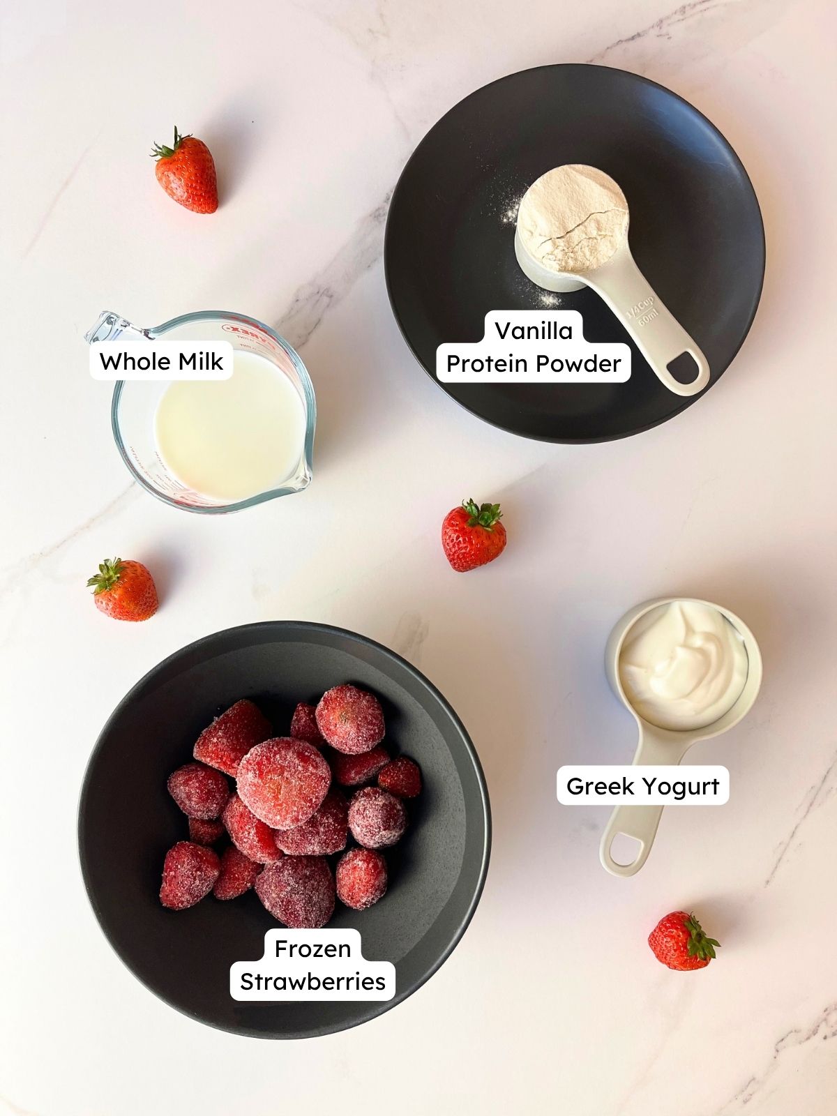 Labeled ingredient overview on a marble surface showing everything needed for the protein strawberry milkshake, including frozen strawberries, whole milk, vanilla protein powder, and Greek yogurt.