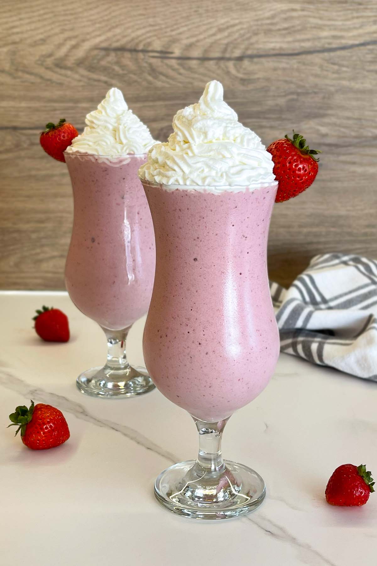 Vertical portrait shot of two finished protein strawberry milkshakes garnished with light whipped cream, surrounded by fresh strawberries on a marble surface.