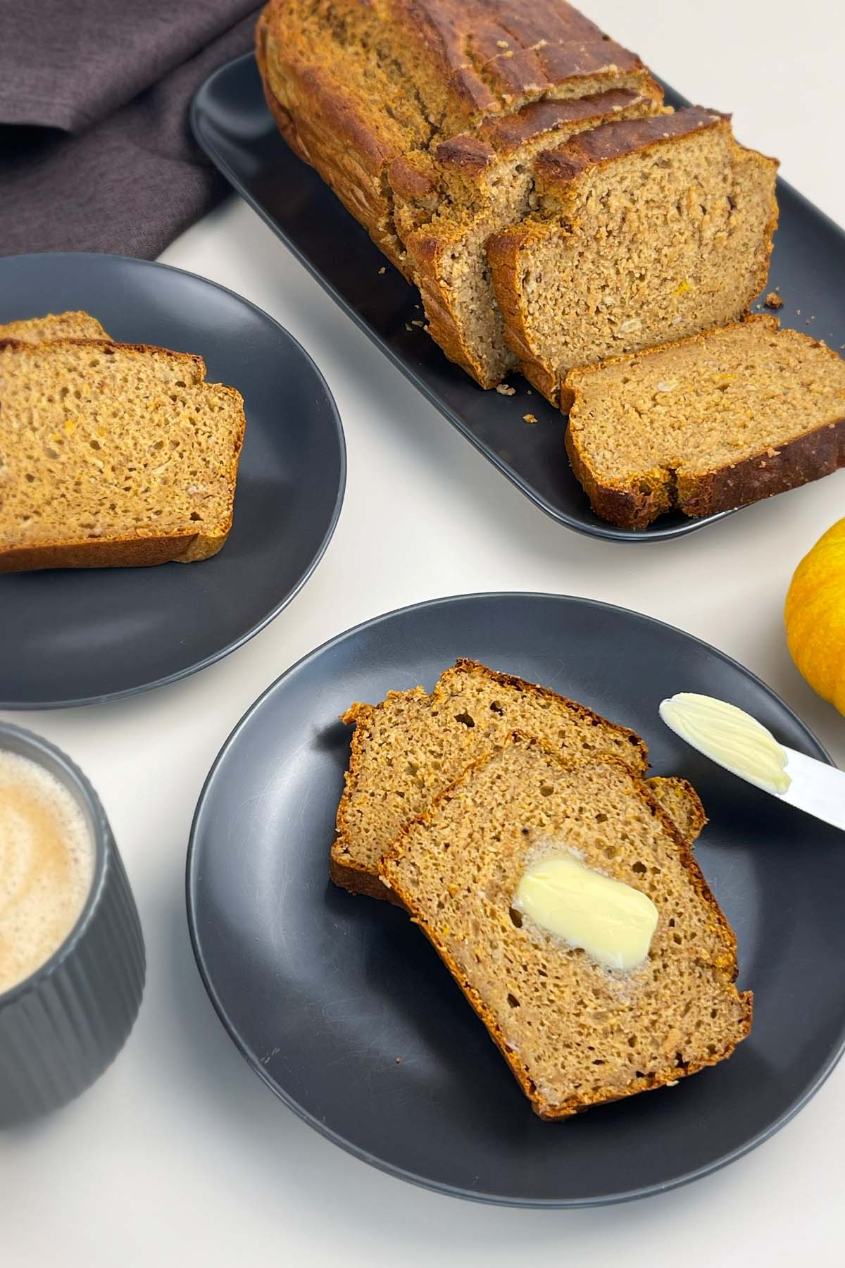 Slices of pumpkin banana bread served on dark gray plates, with one slice spread with a melting pat of butter, next to a cup of coffee.