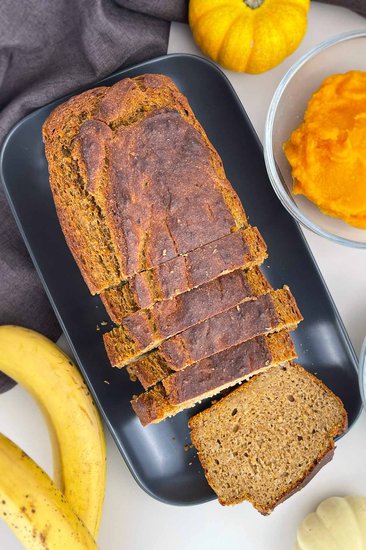 Top-down view of a sliced loaf of pumpkin banana bread on a dark serving platter, surrounded by fresh bananas and pumpkin puree.